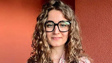 Una donna con lunghi capelli ricci che indossa una camicia fantasia rosa chiaro, in piedi contro un muro rosso testurizzato.