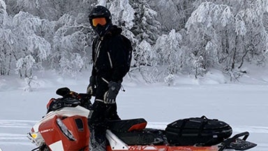 En person klädd i svart vinterutrustning och en hjälm, som står på en röd snöskoter i ett snöigt landskap med frosttäckta träd.
