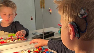 Ett barn som leker med färgglada leksaksklossar på ett bord och tittar i en spegel. En audioprocessor för cochleaimplantat syns på örat.